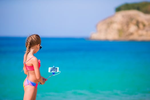Little girl taking selfportrait by cellphone on the beach. Kid enjoying her suumer vacation and making photos background the sea