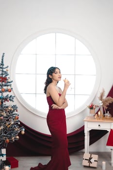 Portrait of a pretty young girl wearing a red gown and drinking wine, smiling at the camera, standing in decorated Christmas living room indoors photo