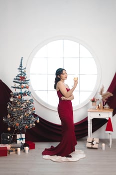 Portrait of a pretty young girl wearing a red gown and drinking wine, smiling at the camera, standing in decorated Christmas living room indoors photo