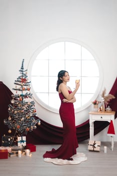 Portrait of a pretty young girl wearing a red gown and drinking wine, smiling at the camera, standing in decorated Christmas living room indoors photo
