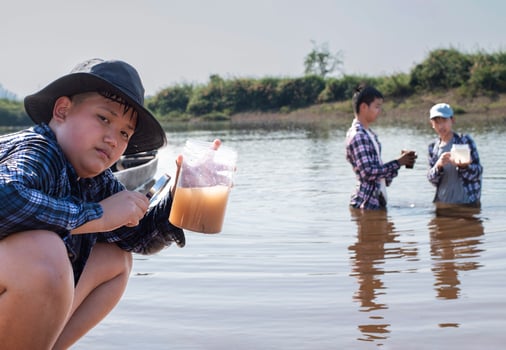 Young asian boy holds transparent tube which has example water inside to do the experiment and ph level measurement as his school project work with his friends behind at the river where he lived. photo