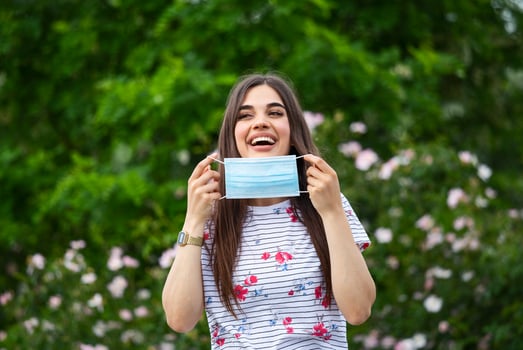 Happy positive girl, young beautiful woman takes off protective sterile medical mask from face outdoors, smiling. Happy end. Pollen allergy. Victory over coronavirus. Pandemic Covid-19. Spring flowers photo