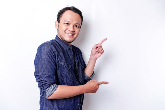 Excited Asian man wearing blue shirt pointing at the copy space beside him, isolated by white background photo