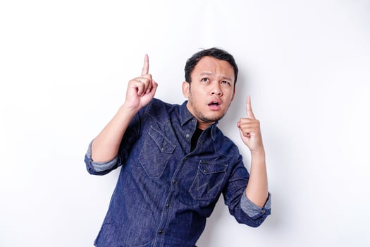 Shocked Asian man wearing blue shirt pointing at the copy space on top of him, isolated by white background photo