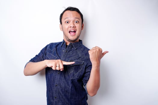 Shocked Asian man wearing blue shirt pointing at the copy space beside him, isolated by white background photo