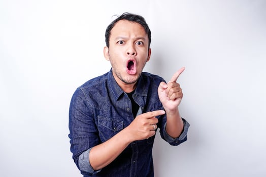 Shocked Asian man wearing blue shirt pointing at the copy space beside him, isolated by white background photo