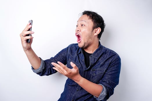 Shocked Asian man wearing blue shirt and holding his phone, isolated by white background photo
