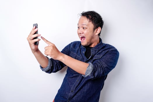 Shocked Asian man wearing blue shirt and holding his phone, isolated by white background photo