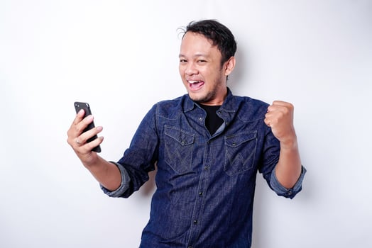 A young Asian man with a happy successful expression wearing blue shirt and holding his phone, isolated by white background photo