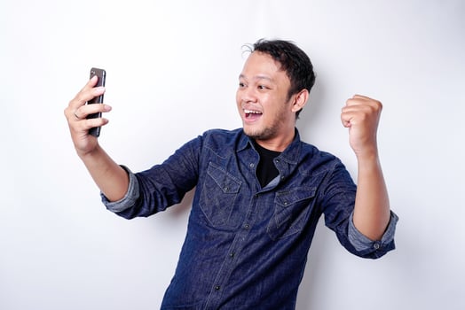 A young Asian man with a happy successful expression wearing blue shirt and holding his phone, isolated by white background photo
