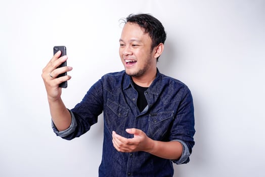 Excited Asian man wearing blue shirt pointing at the copy space beside him while holding his phone, isolated by white background photo