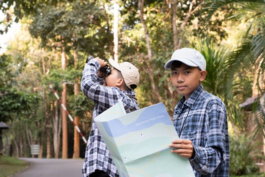 Asian young teen boys hold national park map, reading details of birdwatching before using their binoculars to watch the bird which sitting on brunches and flying in the sky, summer vacation concept. photo