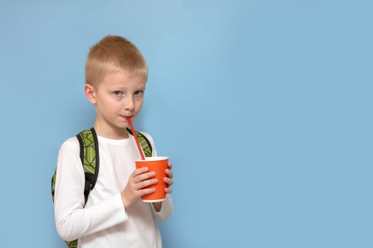 boy with a backpack on his back holds a glass in his hands and drinks through a straw on a blue background with copy space photo