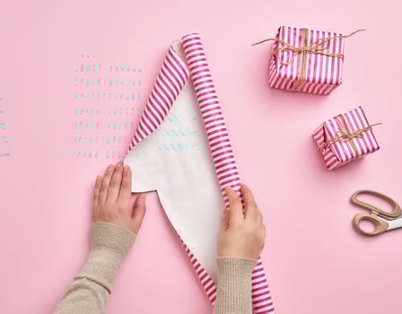 two female hands unfold a roll of wrapping paper, next to two gifts, pink background photo