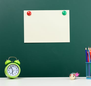 blank sheet of paper affixed on a green chalk board, alarm clock and multi-colored pencils. Back to school photo