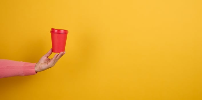hand holds paper cardboard red cup for coffee, yellow background. Eco-friendly tableware, copy space photo