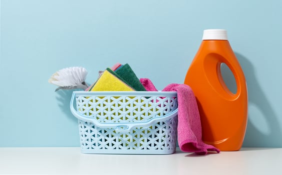 basket with spoges and a brush, next to a plastic orange bottle with liquid detergent on a white table photo