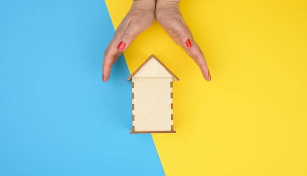 two female hands folded to each other over a wooden miniature model house on a yellow-blue background. Real estate insurance concept photo