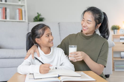 Asian young little girl learn at home. Do homework with kind mother help, encourage for exam. Mom pass on a glass of milk to daughter. Girl happy Homeschool. Mom teach and advice education together. photo