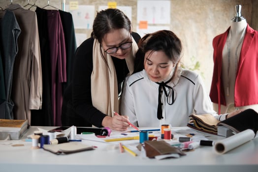 Asian middle-aged female fashion designer teaches a young teen trainee tailor in studio with colorful thread and sewing fabric for dress design collection ideas, professional boutique small business. photo
