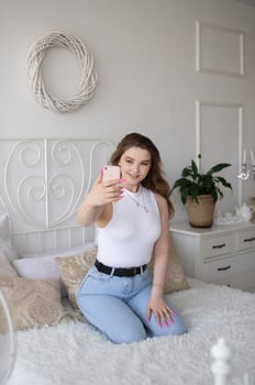 A cute girl sits on a white bed with a phone and takes a selfie photo