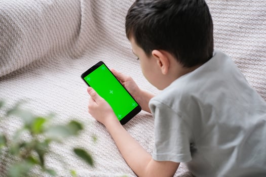 The boy looks at the screen of the phone with a chrome key indoors. An unrecognizable child uses a smartphone with a green screen to watch a video. photo