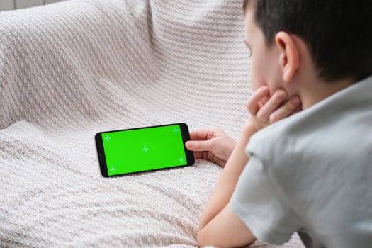 The boy looks at the screen of the phone with a chrome key indoors. An unrecognizable child uses a smartphone with a green screen to watch a video. photo