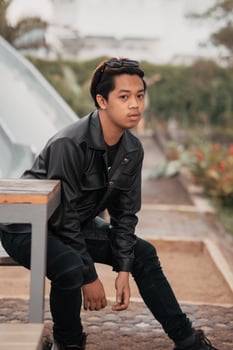 an Asian man is sitting on a wooden chair while wearing a black leather shirt and jacket while stylish with sunglasses photo