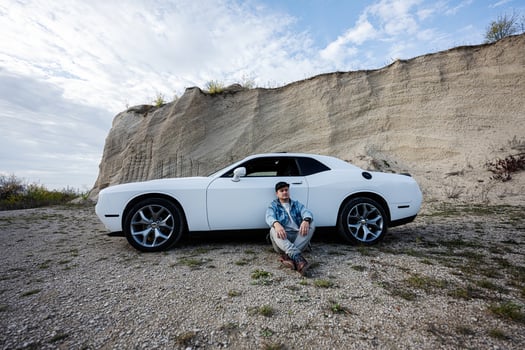 Handsome man in jeans jacket and cap sitting near his white muscle car in career. photo