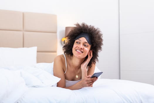 Happy girl using a mobile phone lying on the bed at home. Beautiful young smiling woman lying in her bed with sleeping eye mask and using a phone in her bedroom. photo