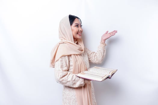 Young Asian Muslim woman smiling while pointing to copy space beside her and holding the Quran photo