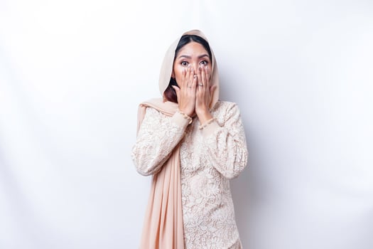 Oops Surprised young Asian Muslim woman covering mouth with hands and staring at camera while standing against white background photo