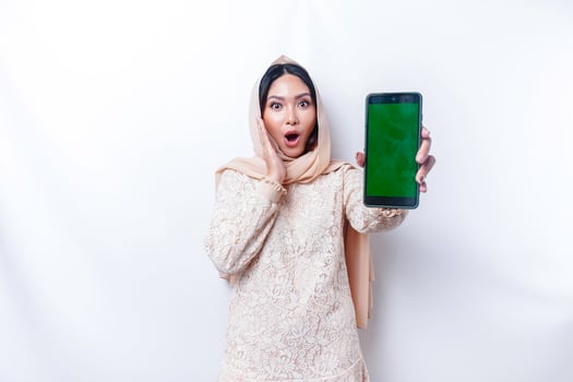 Shocked Asian woman wearing hijab, showing copy space on her phone screen, isolated by white background photo