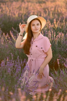 A cheerful young girl in a pink dress and a straw hat in her hands stands among lavender bushes. Sunset. photo