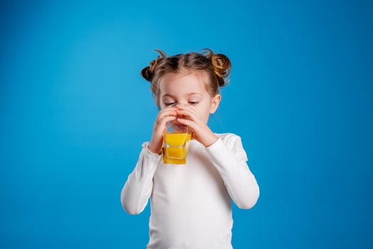 little girl with a funny hairstyle in a white T-shirt drinks juice from a glass. blue background. useful products for children. space for text. High quality photo