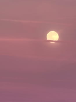 A hazy yellow moon peeking out through the pink and purple sunset sky photo