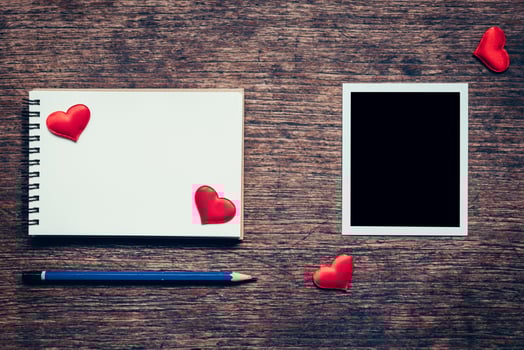 Blank photo frame, notebook, pencil and red heart on wood table background.