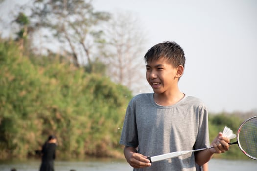 Asian boys hold badminton shuttlecock and racket, standing and playing beside the river bank in their local river during their weekend holiday, soft and selective focus on front boy in white shirt. photo
