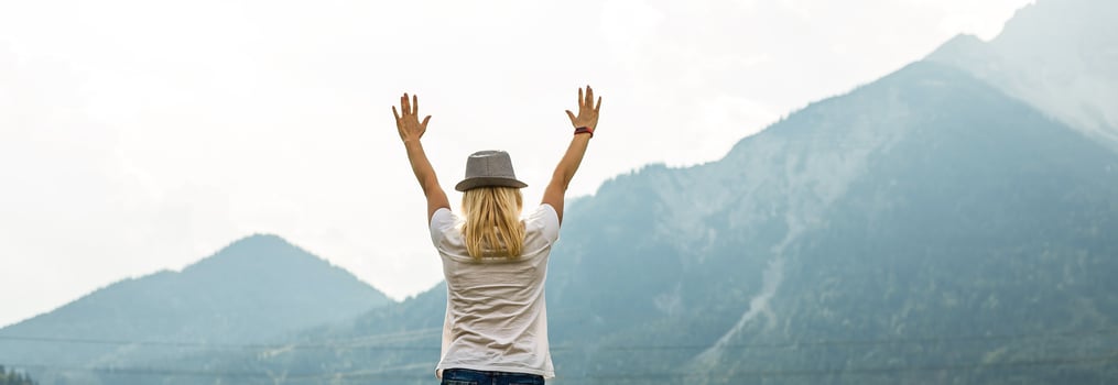 Young woman with backpack standing on cliff's edge and looking to a sky with raised hands photo