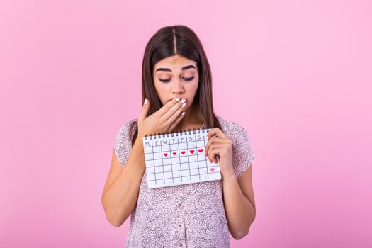Portrait of a beautiful young girl holding her menstruation calendar and covering hear face isolated over pink background. Period calendar photo