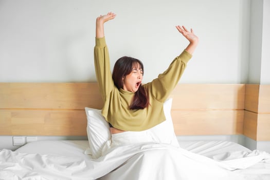 Cute Asian woman stretching out and yawning while sitting on her bed in bright bedroom photo