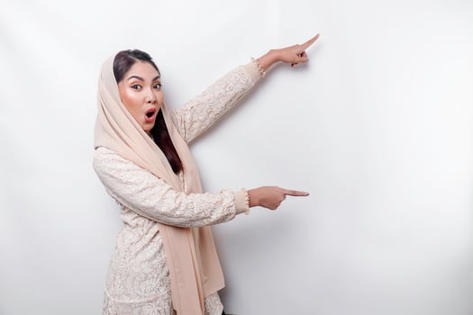 Shocked Asian Muslim woman pointing at the copy space on beside her, isolated by white background photo