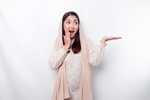 Shocked Asian Muslim woman pointing at the copy space on beside her, isolated by white background photo