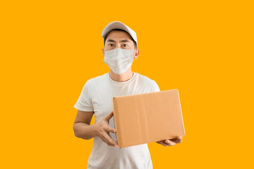 Young Asian delivery man wearing cap and white blank t-shirt holding parcel post box isolated on yellow background. express delivery service concept. photo