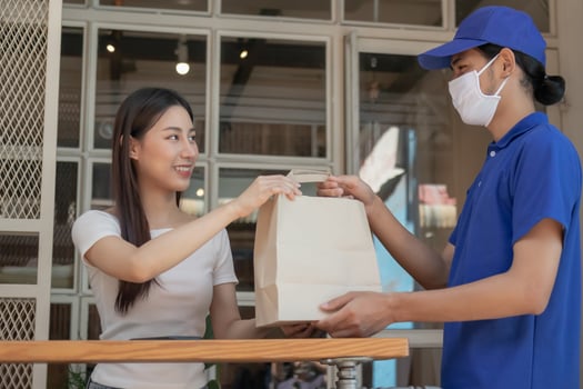 Smile, Happy asian young woman or girl  wearing face mask protect to pandemic of covid-19 and received paper bag from delivery man, messenger in uniform while lockdown, quarantine at front door home. photo