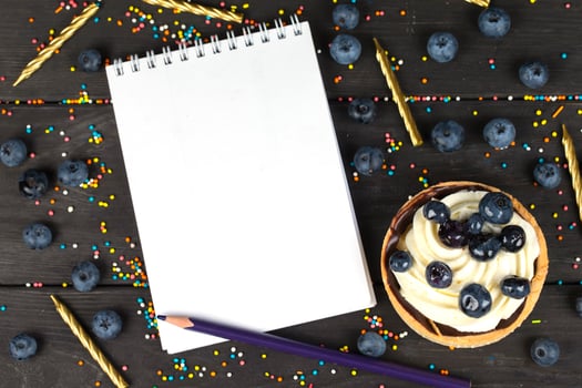 On a wooden board lies a cake with cream and blueberries and a white notebook with blank pages. photo