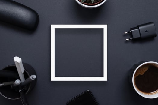 White square paper frame on black office desk surrounded by coffee mug, charger, plant, pens and phone. Copy space in the middle. Top view flat lay photo
