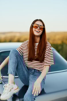 A fashionable woman smiles sweetly in stylish sunglasses, a striped t-shirt and jeans, sits on the trunk of a car and looks at the beautiful nature of autumn. Travel lifestyle photo