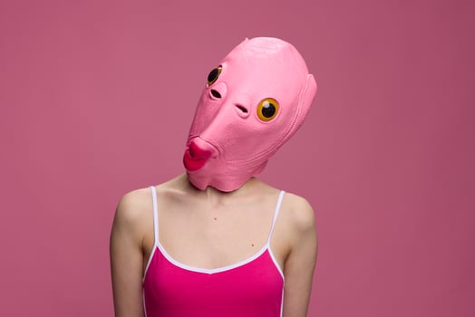 Woman wearing a pink fish head Halloween mask stands and looks at the camera against a pink background photo
