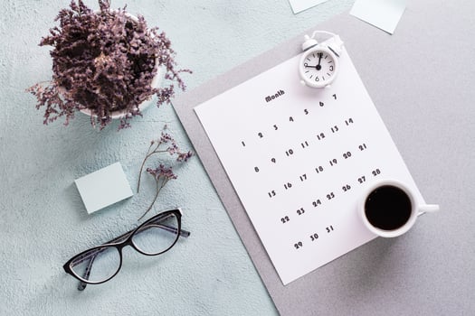 Monthly calendar sheet, coffee cup, alarm clock, glasses and a bouquet of flowers on the table. Time planning and organization photo